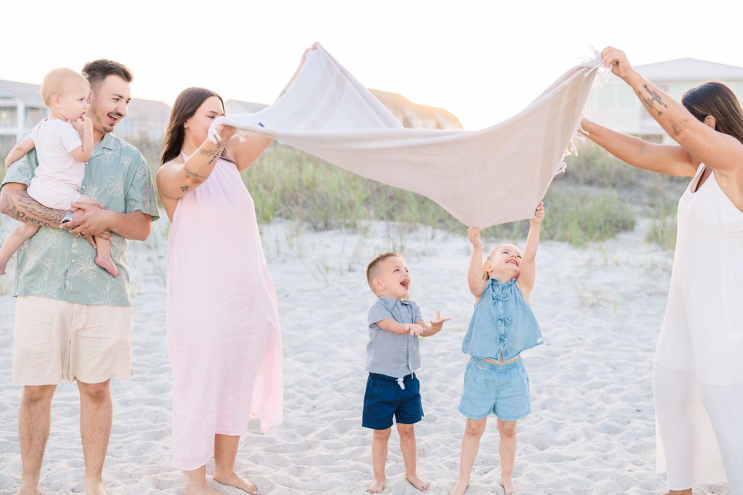 Family playing with blanket on sandy beach at sunset, two adults and three young children laughing outdoors.
