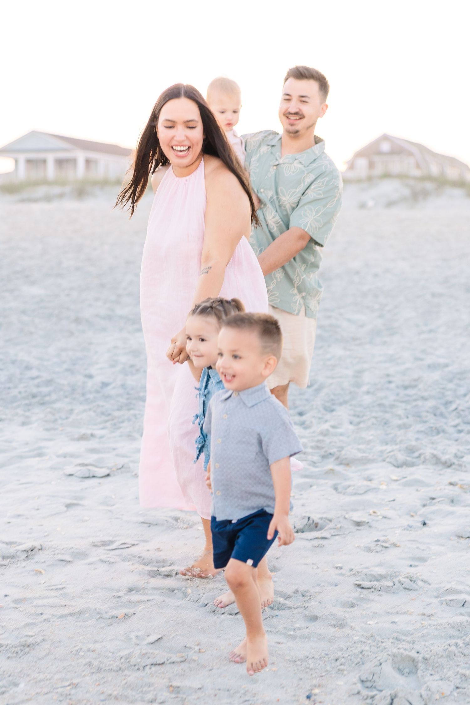 Family laughing and playing on sandy beach at sunset, mother in pink dress with three young children and father in green shirt.