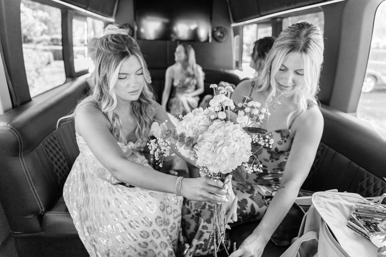 Two women in formal dresses handle flowers inside a vintage car in black and white.