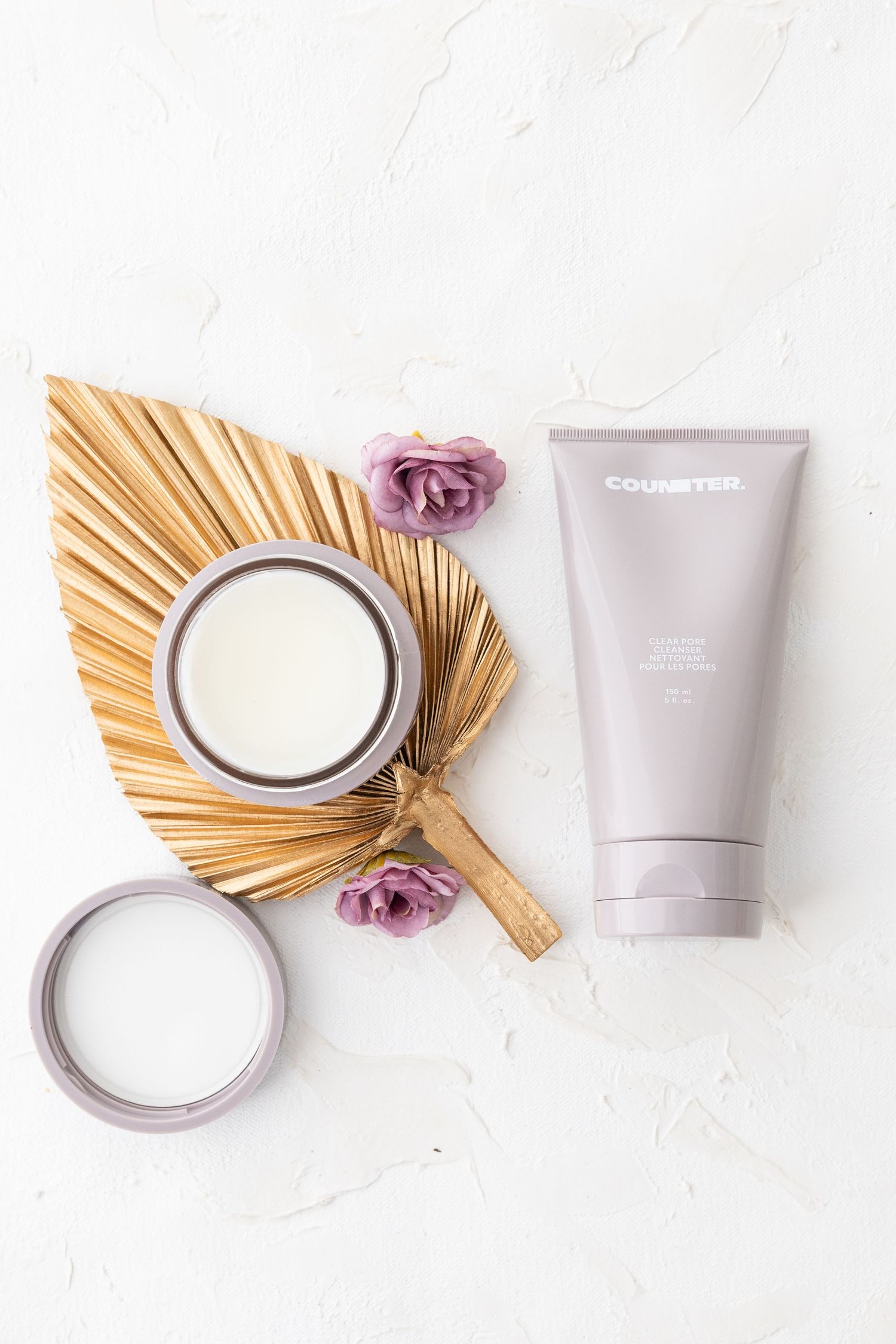 Elegant cosmetic products arranged with dried palm fronds and purple roses on white surface.