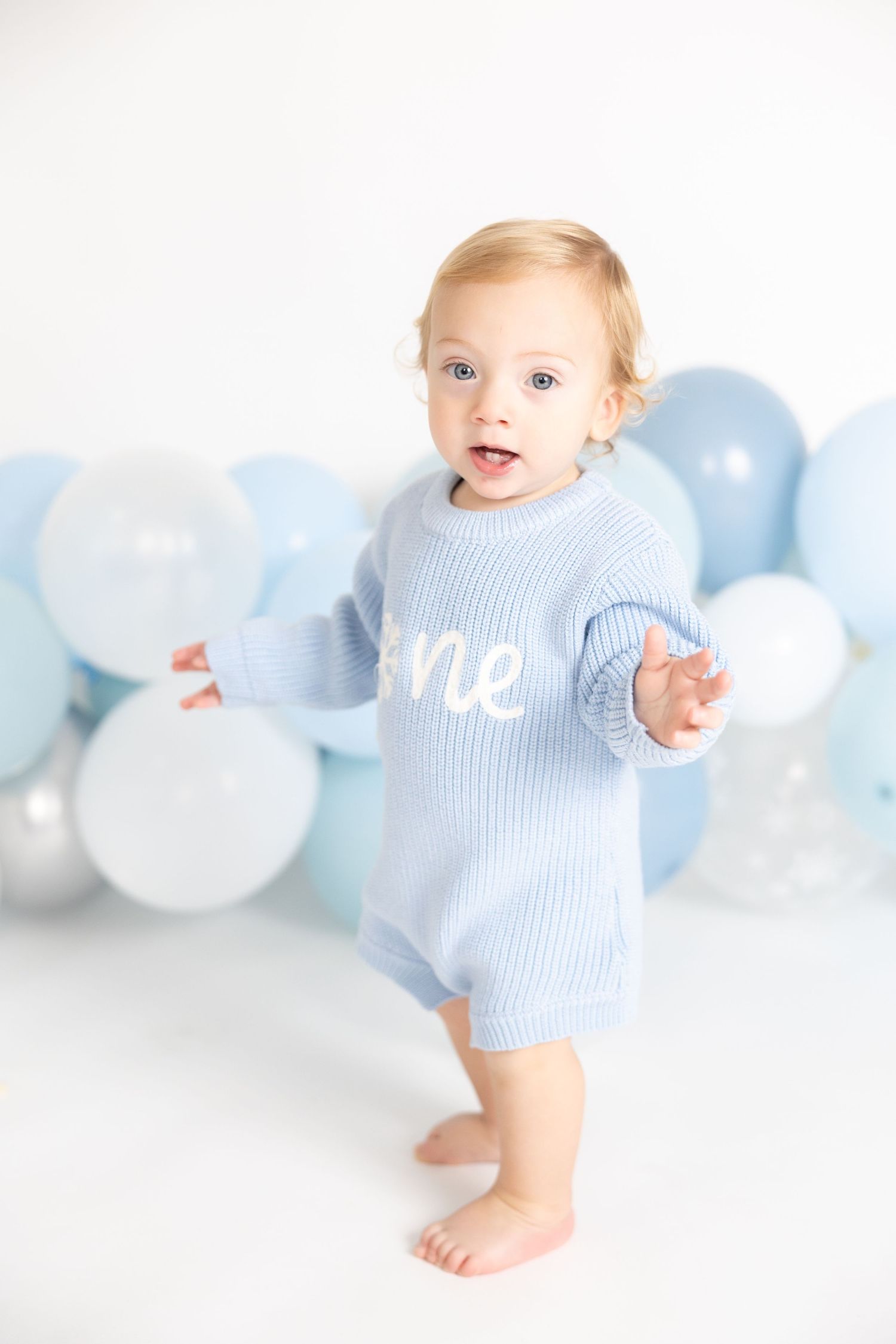 Baby wearing light blue outfit stands near balloon display in white studio setting.