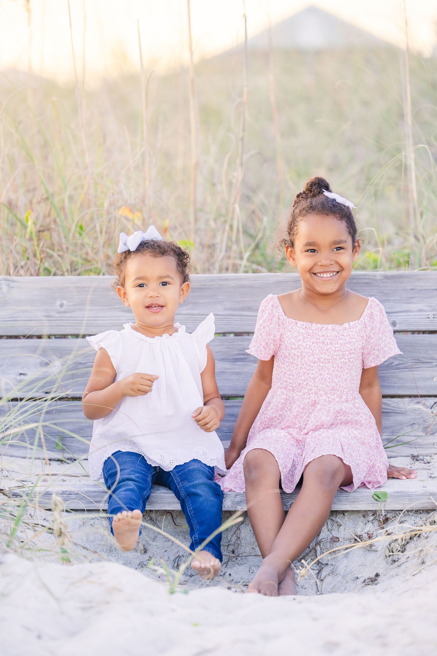 Two children in coordinated pink and white outfits sit together on a wooden bench at the beach.