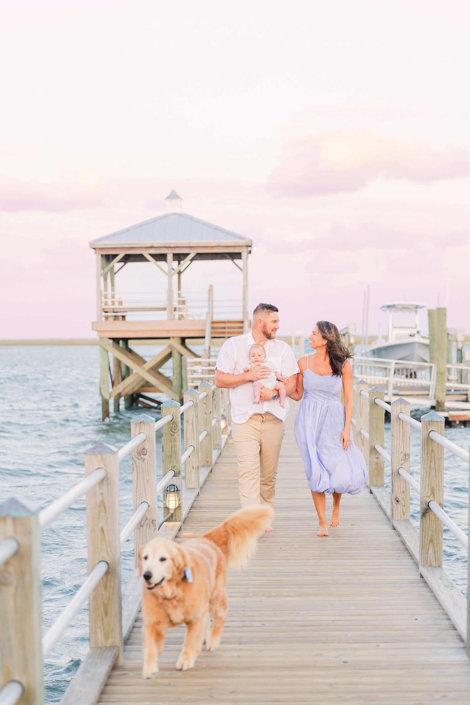 A couple walks their golden retriever on a coastal pier with a white gazebo in the background.