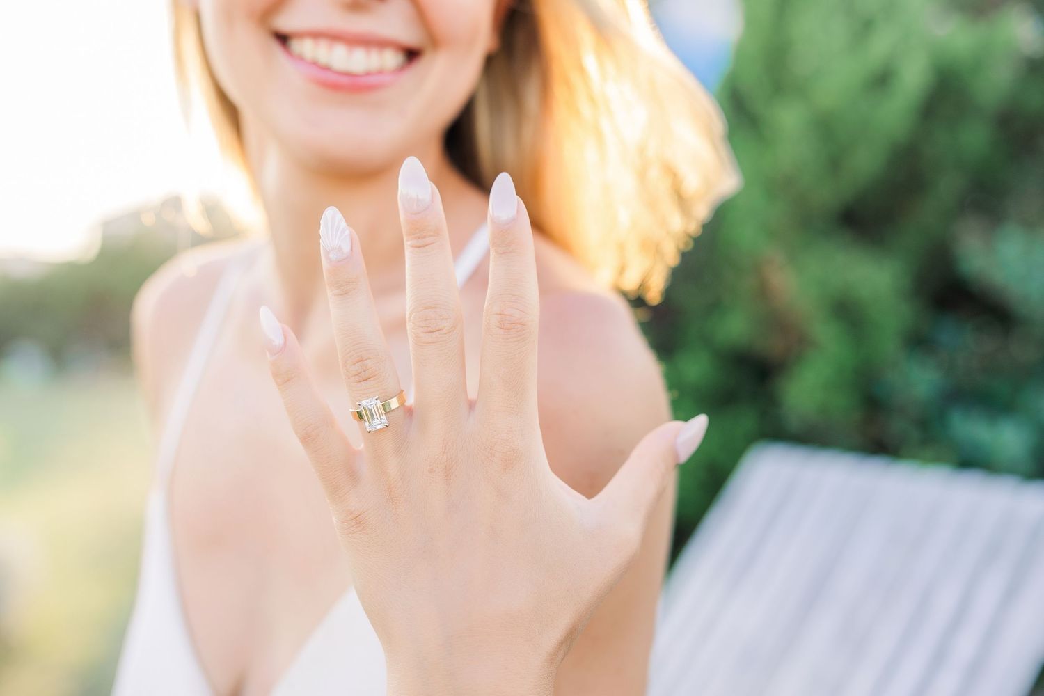 Smiling bride-to-be showing off her stunning yellow gold engagement ring with long nude nails outdoors.