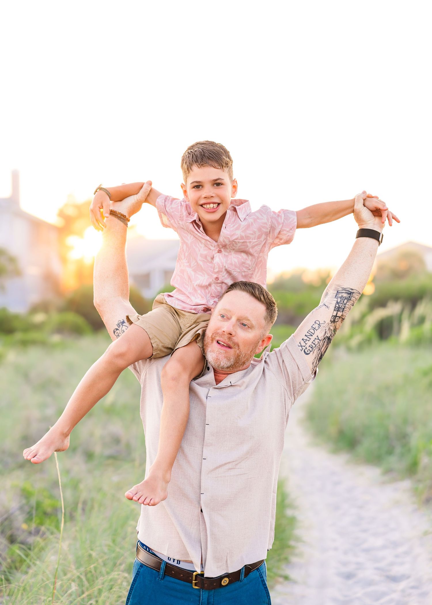 Man with tattoos holds smiling boy on shoulders at sunset beach path.