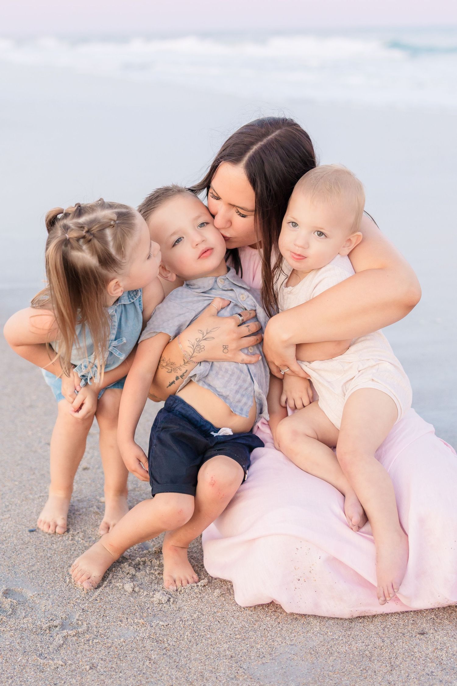 Mother sitting on beach hugging three young children close, with ocean waves in the background.