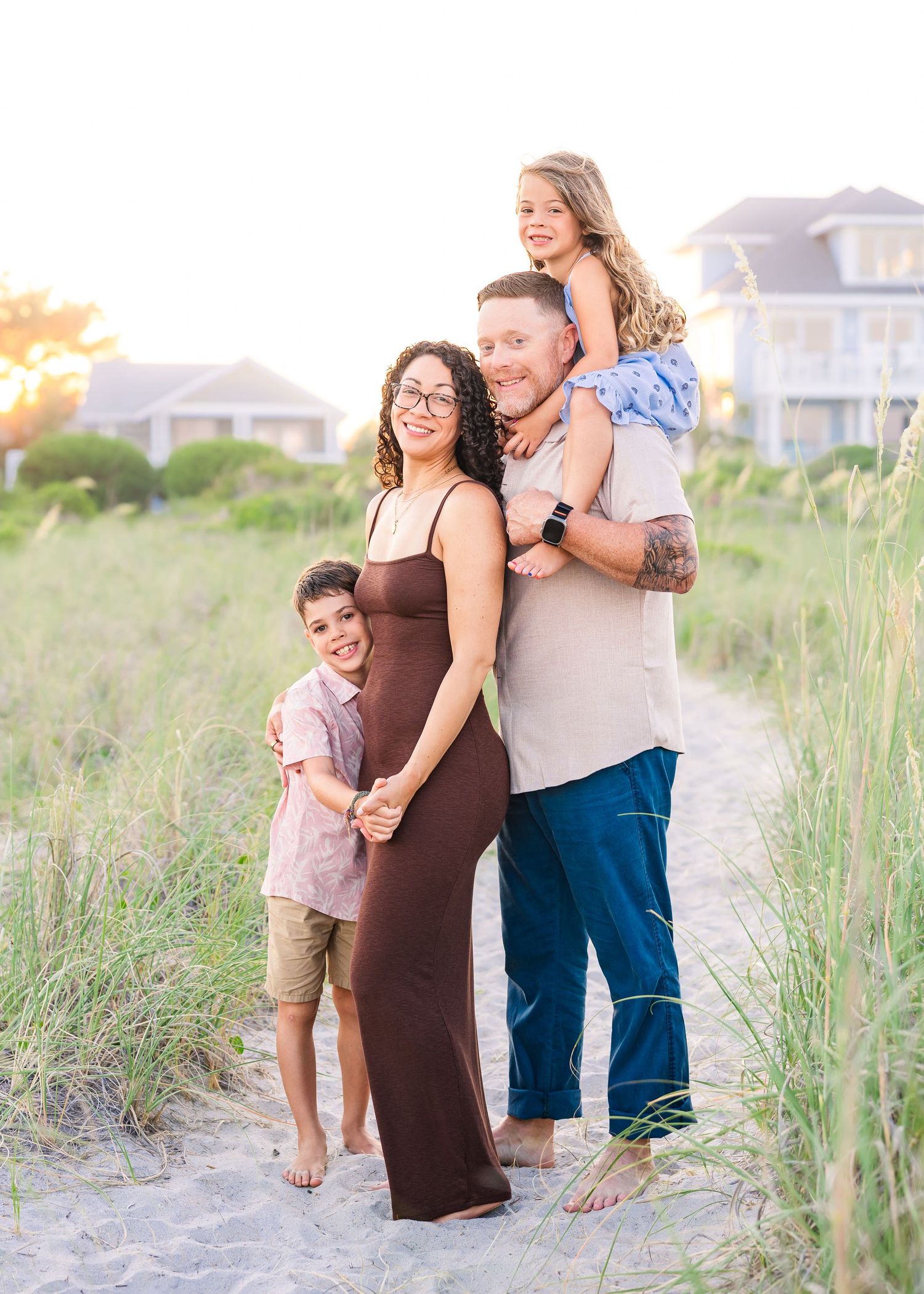 Family of four poses on a sandy beach path at golden hour, with two kids and parents smiling warmly.