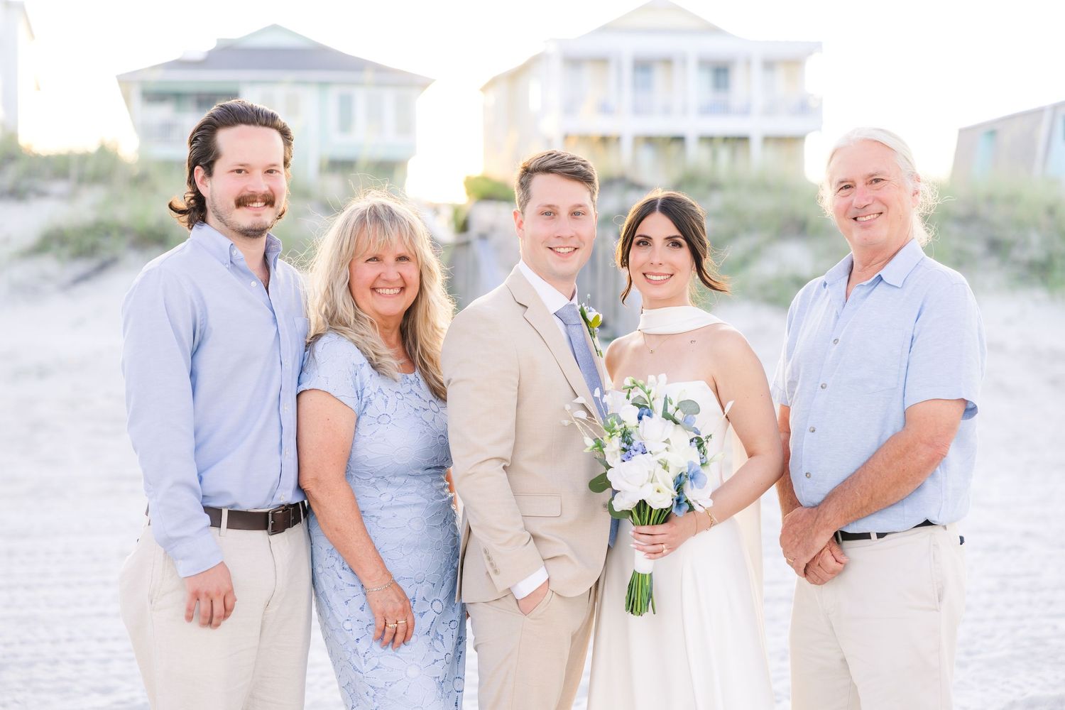 Bride and groom pose with family on beach, bride holding white floral bouquet at sunset in Carolina Beach near Wilmington, NC.