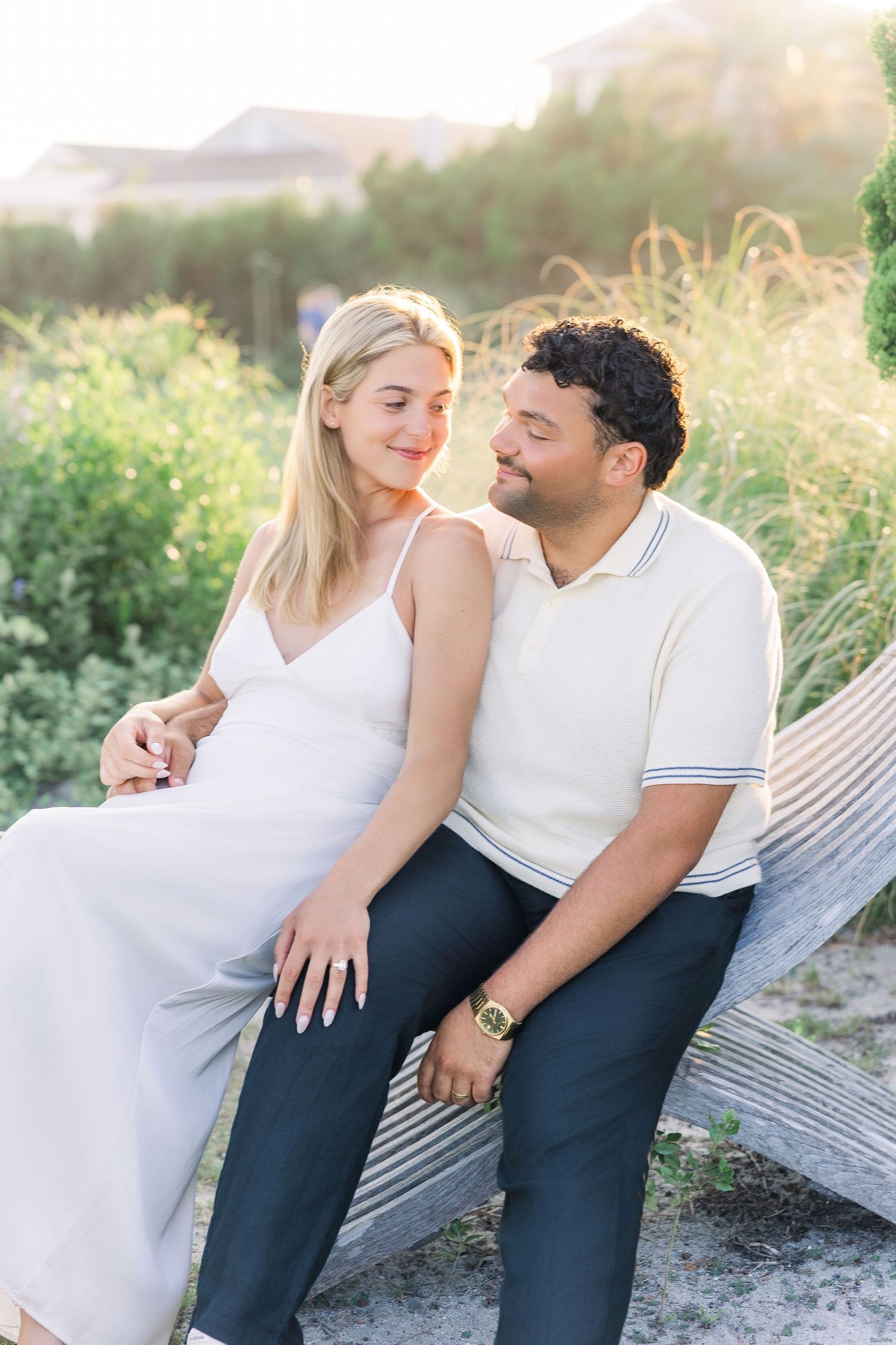 Couple sitting together on beach dunes at golden hour, woman in white dress, man in cream polo shirt.