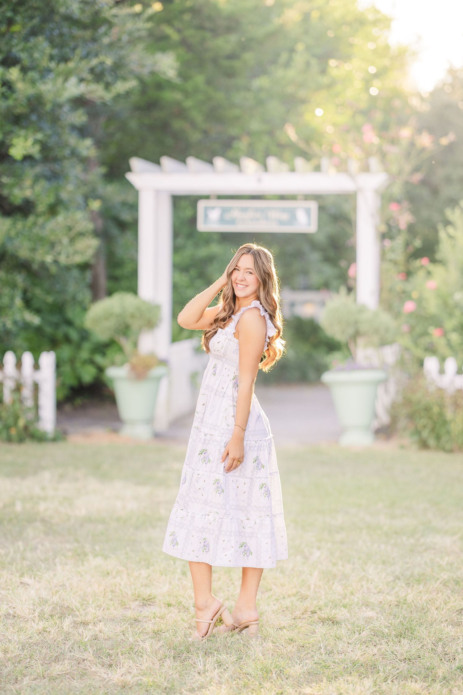 Young girl in white lace dress twirls playfully in a sunny garden setting with white fence and arbor in background.
