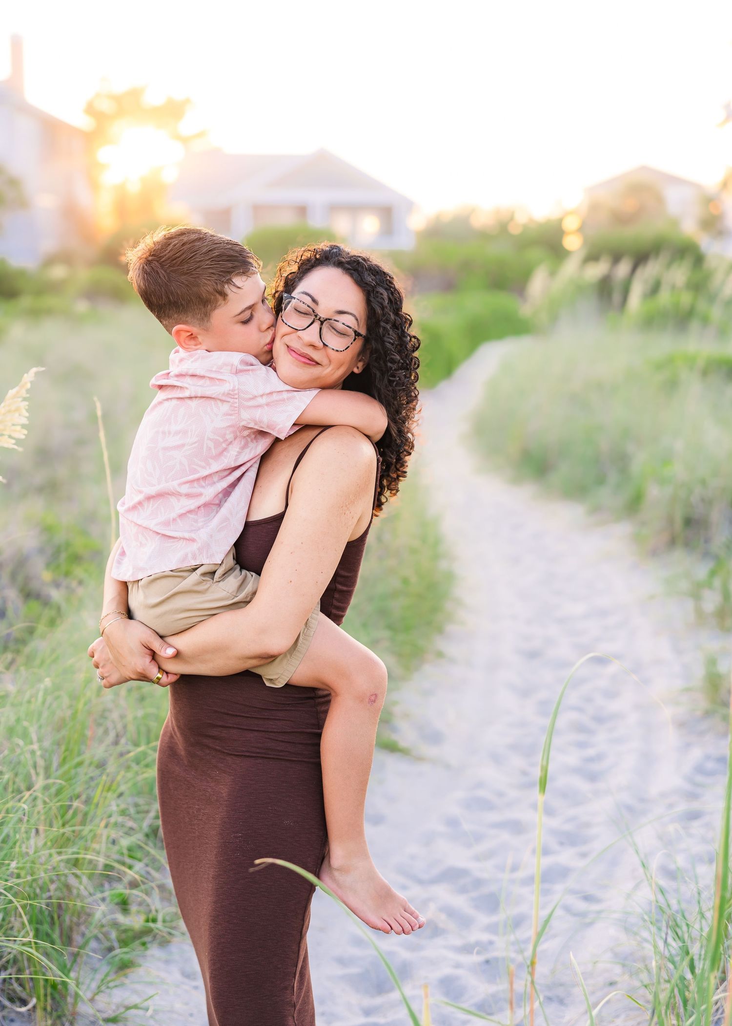 A mother with curly hair and glasses holds her young son on a sandy beach path at golden hour.