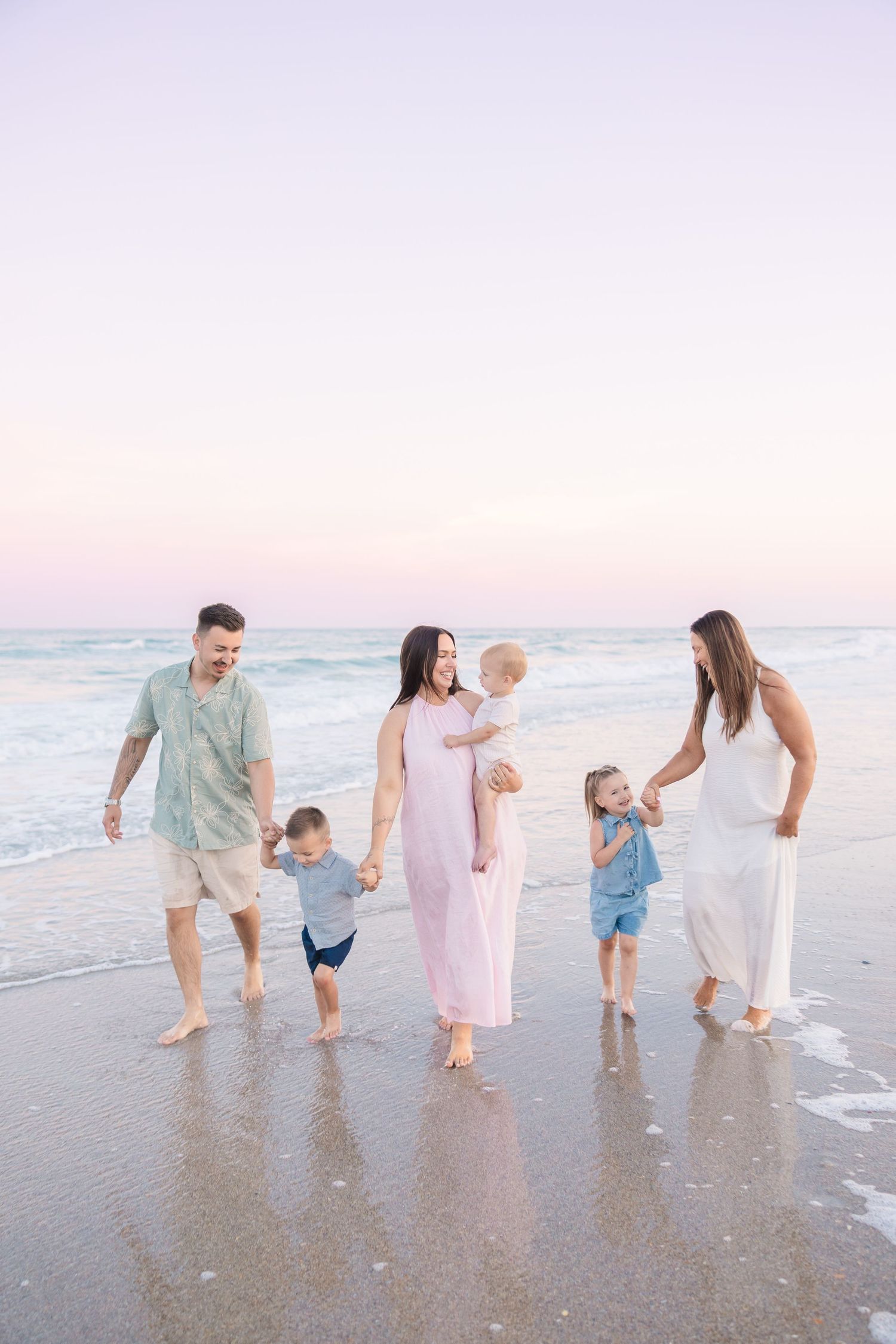 Extended family of six walks along shallow beach shoreline at sunset, children playing in water.