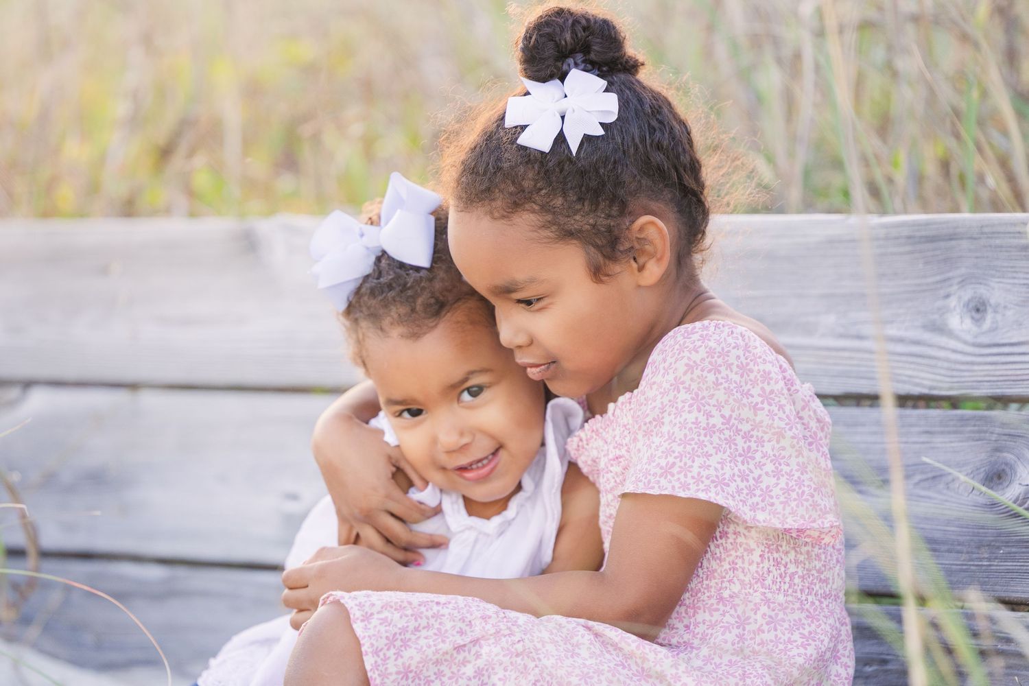 Two children in matching white bows share a tender embrace while sitting on a wooden bench outdoors.