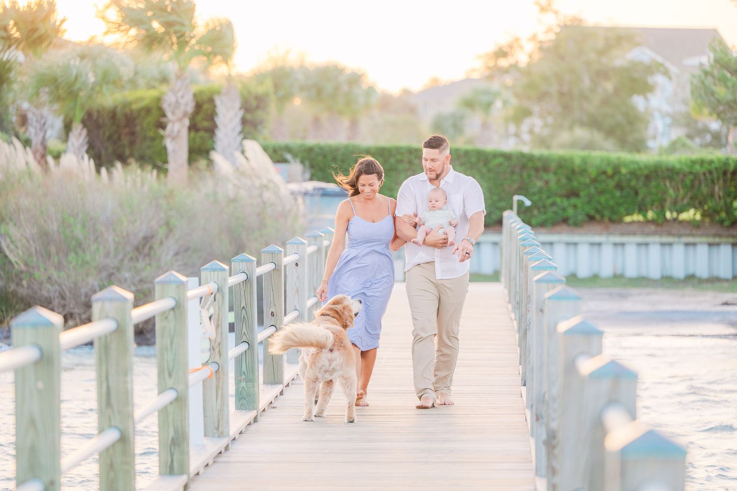 A couple walks their golden retriever along a coastal boardwalk during golden hour.