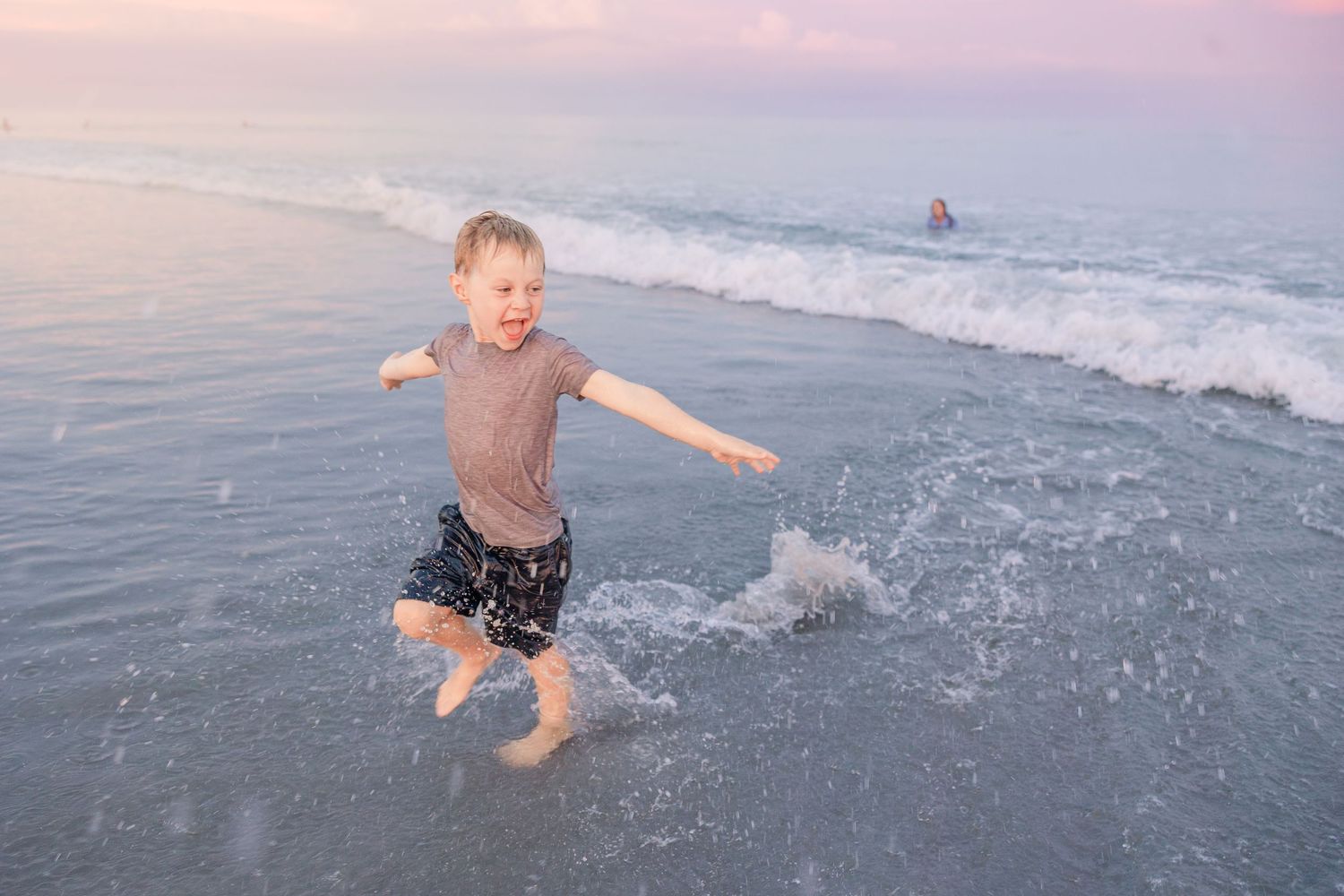 A joyful child splashing in the ocean waves during a pink sunset.
