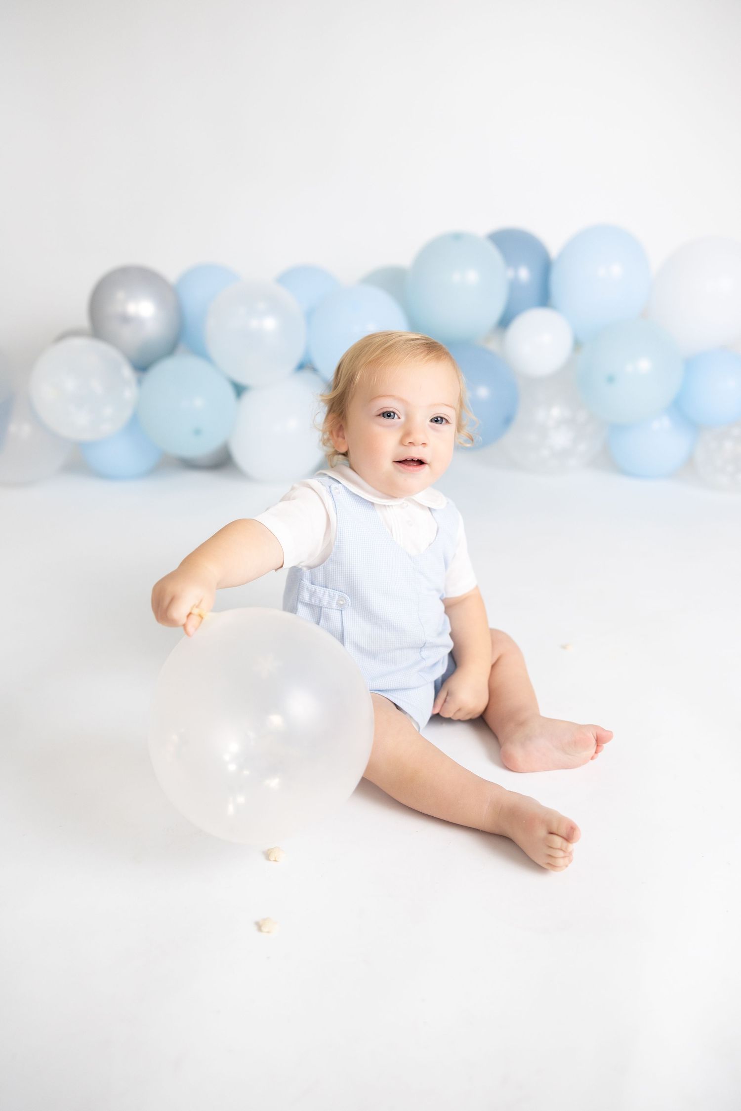 Baby in white outfit poses against backdrop of soft blue and white balloons in minimalist studio setting.