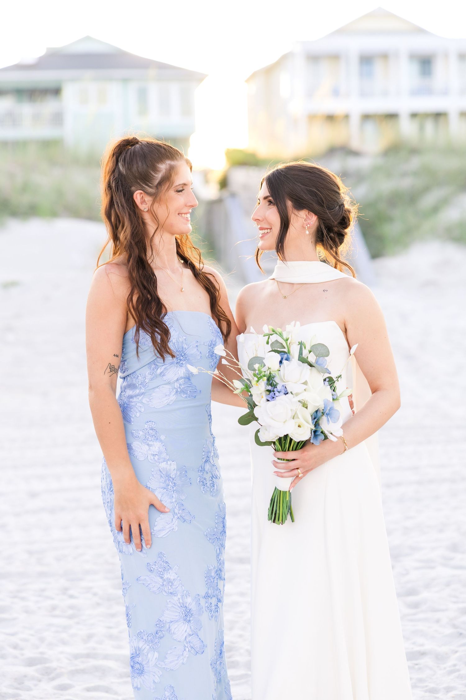 Bride in white gown with blue floral bouquet smiles with bridesmaid in light blue floral dress on beach in Carolina Beach near Wilmington