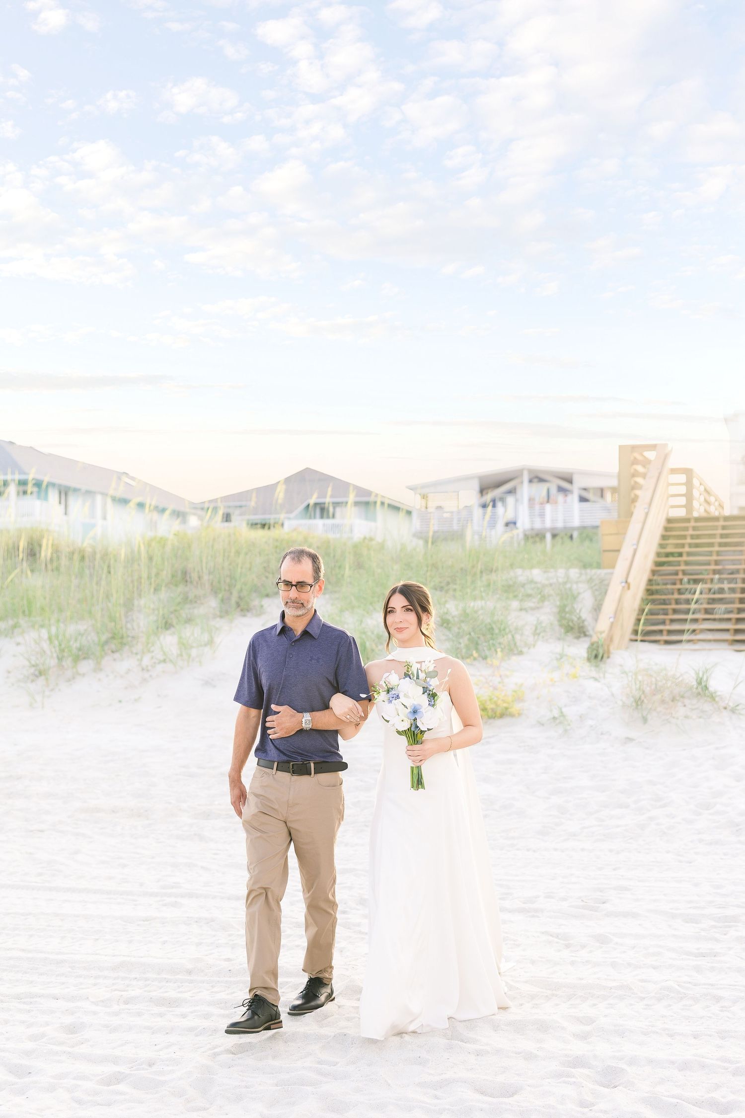 Bride in white gown holds blue and white bouquet standing with suited man on sandy beach at sunset in Carolina Beach near Wilmington, NC.