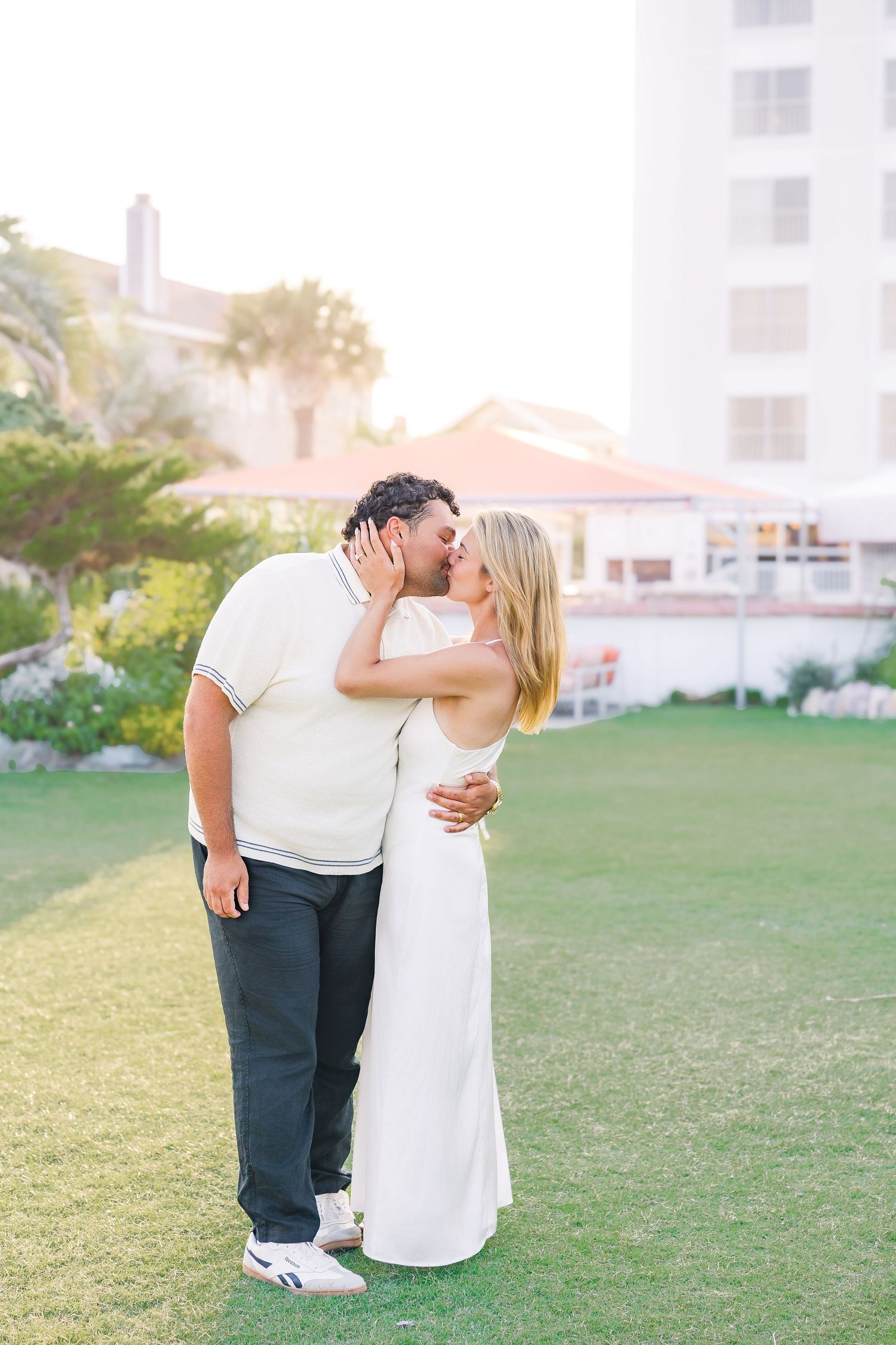 Couple sharing a romantic kiss on a green lawn with a white building and palm trees in the background.