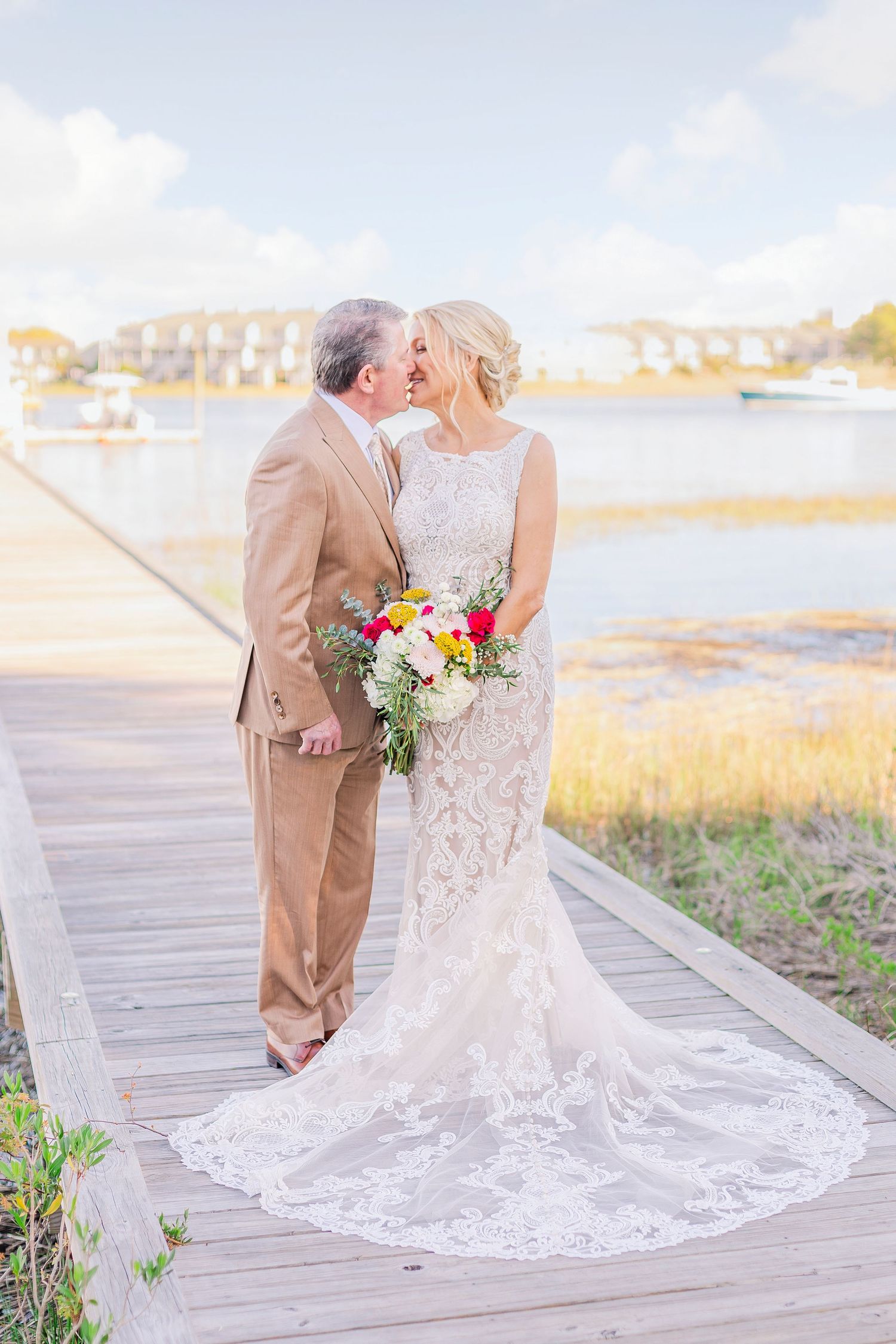 A romantic wedding couple poses on a wooden boardwalk with a flowing lace dress train and colorful bouquet.