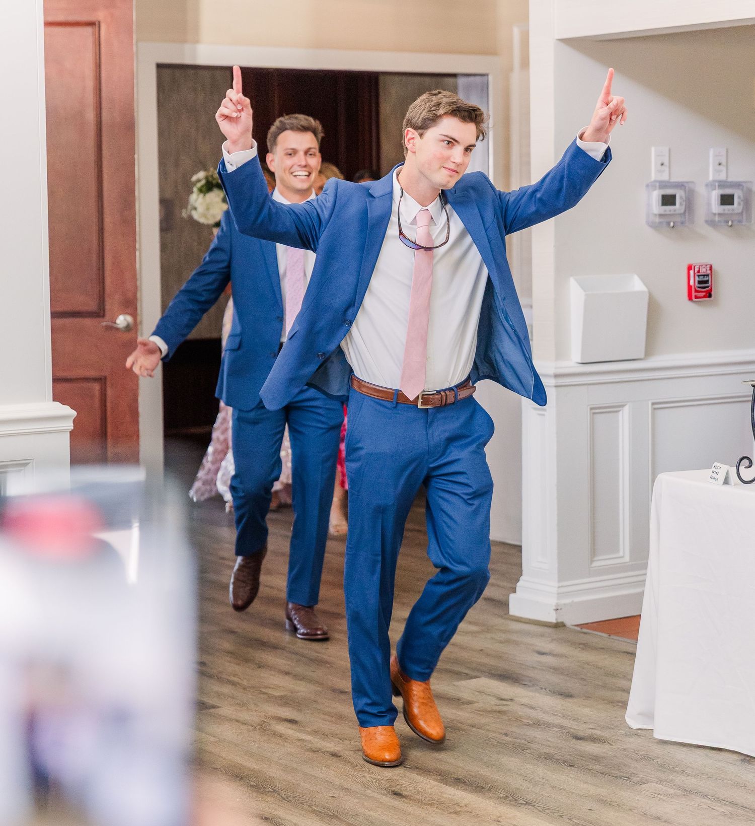 Wedding party members in blue suits dance and celebrate in an indoor venue.