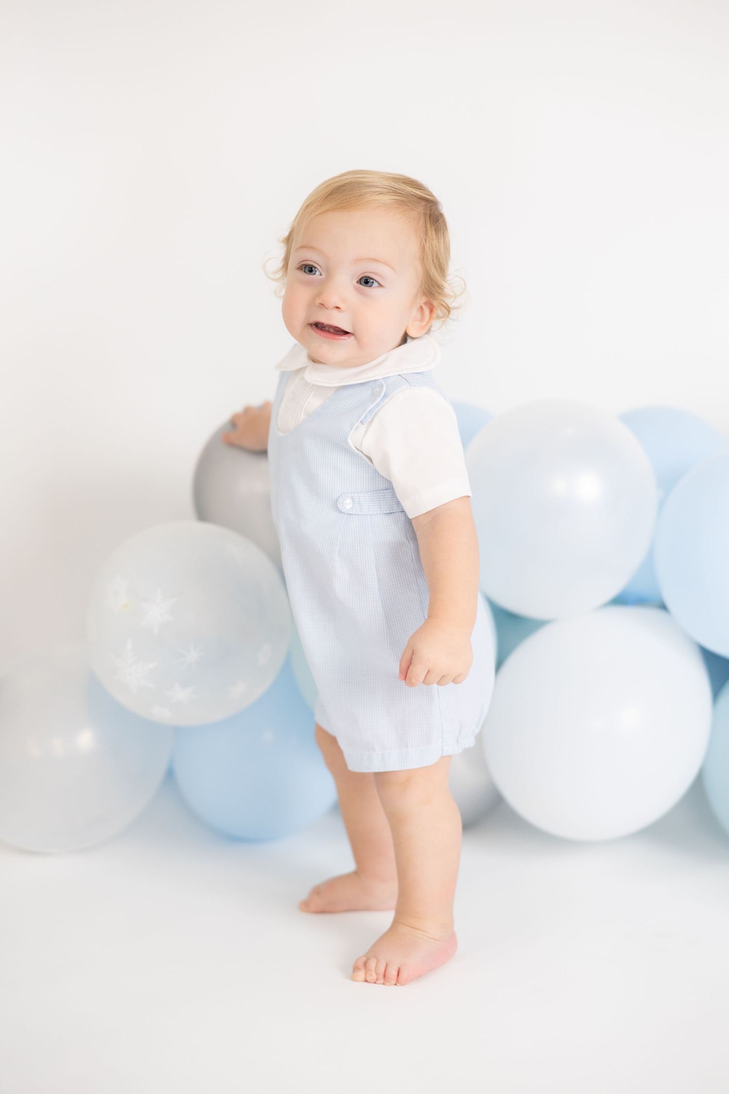 Baby standing against white wall with blue and silver balloon arrangement.