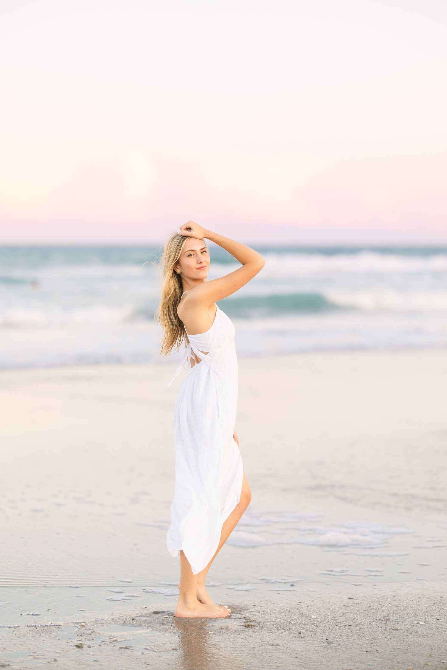 Graceful pose in white dress on beach during soft pink sunset.
