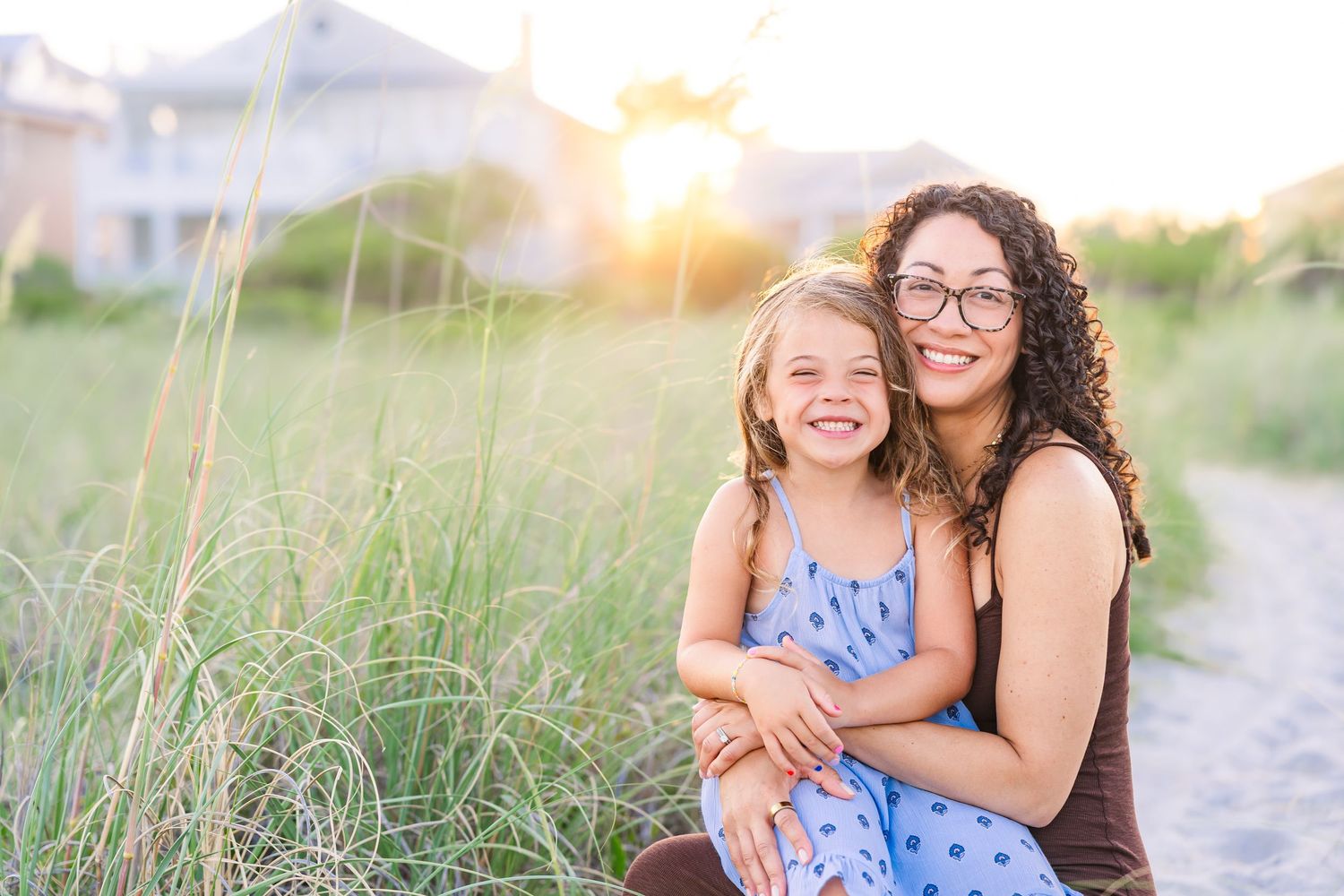 A woman and young girl smile together on a sandy beach path at golden hour sunset.
