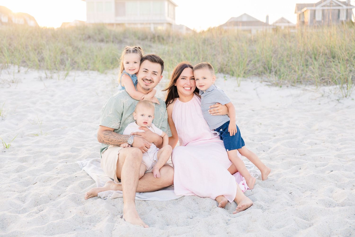 A smiling family of five sits together on sandy beach at golden hour.