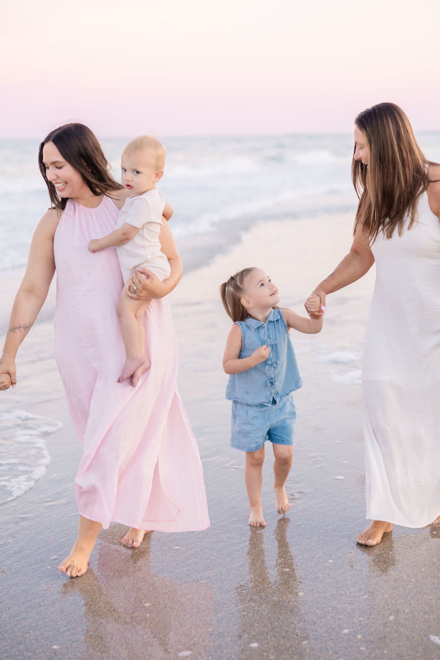 Two women and two young children walking together along a beach at sunset with ocean waves.