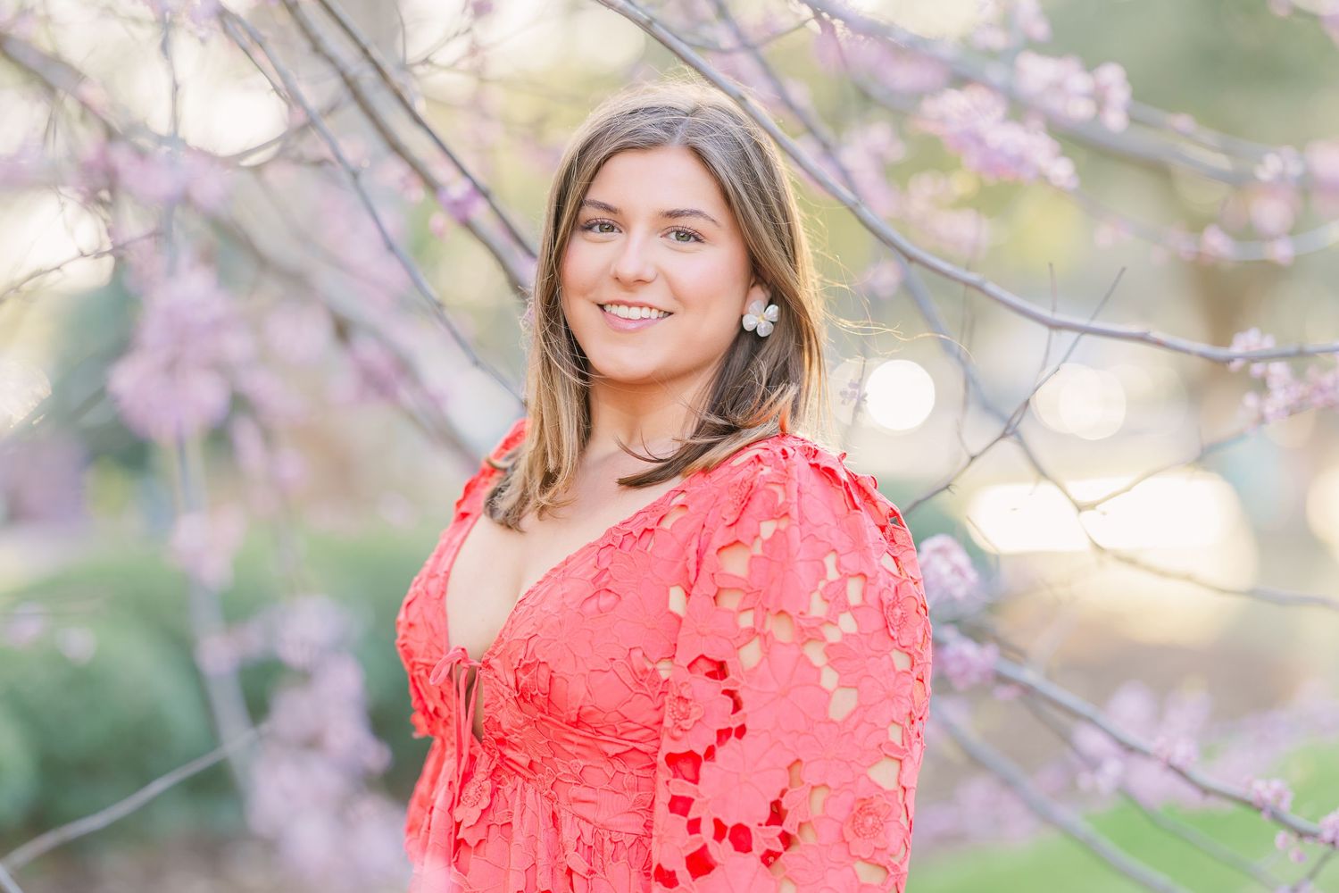 A person in a bright coral dress poses among blooming cherry blossom trees in springtime.