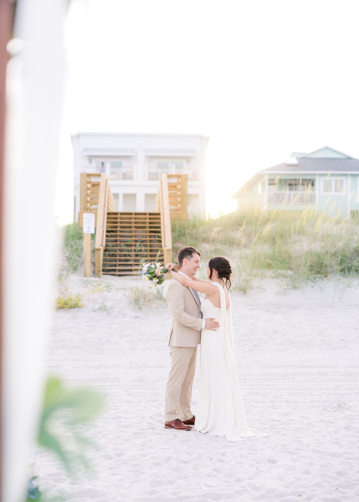 Bride and groom share a romantic moment on a sandy beach with coastal homes and dunes in soft golden light in Carolina Beach near Wilmington