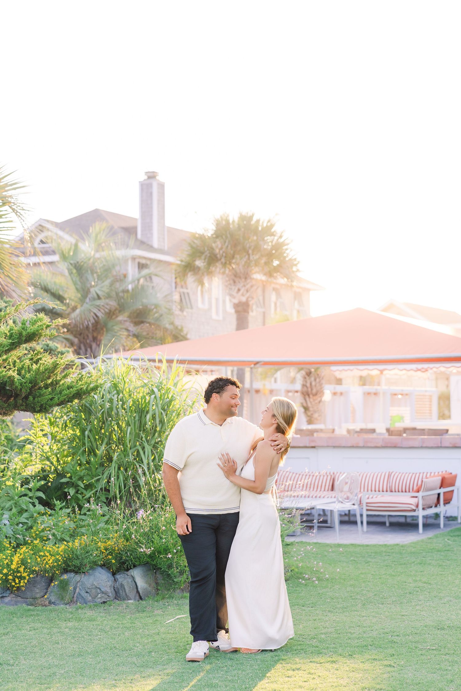 Couple sharing a romantic moment on green lawn with tropical resort backdrop in warm golden sunset light.