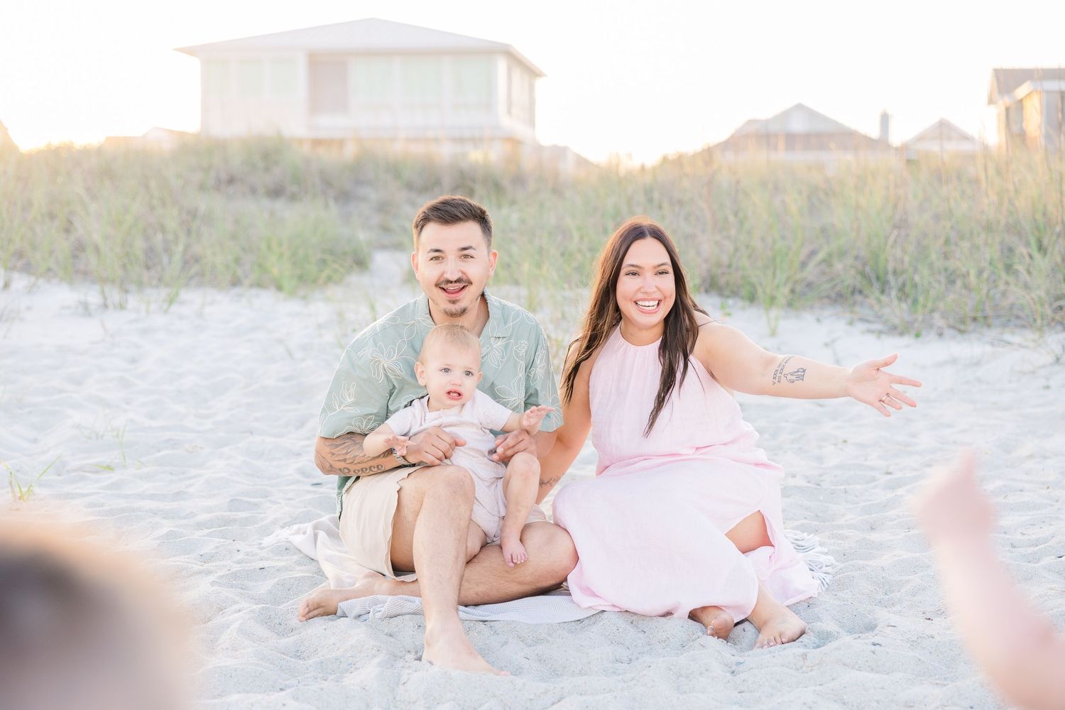 Happy couple sitting on sandy beach with their laughing baby at golden hour.