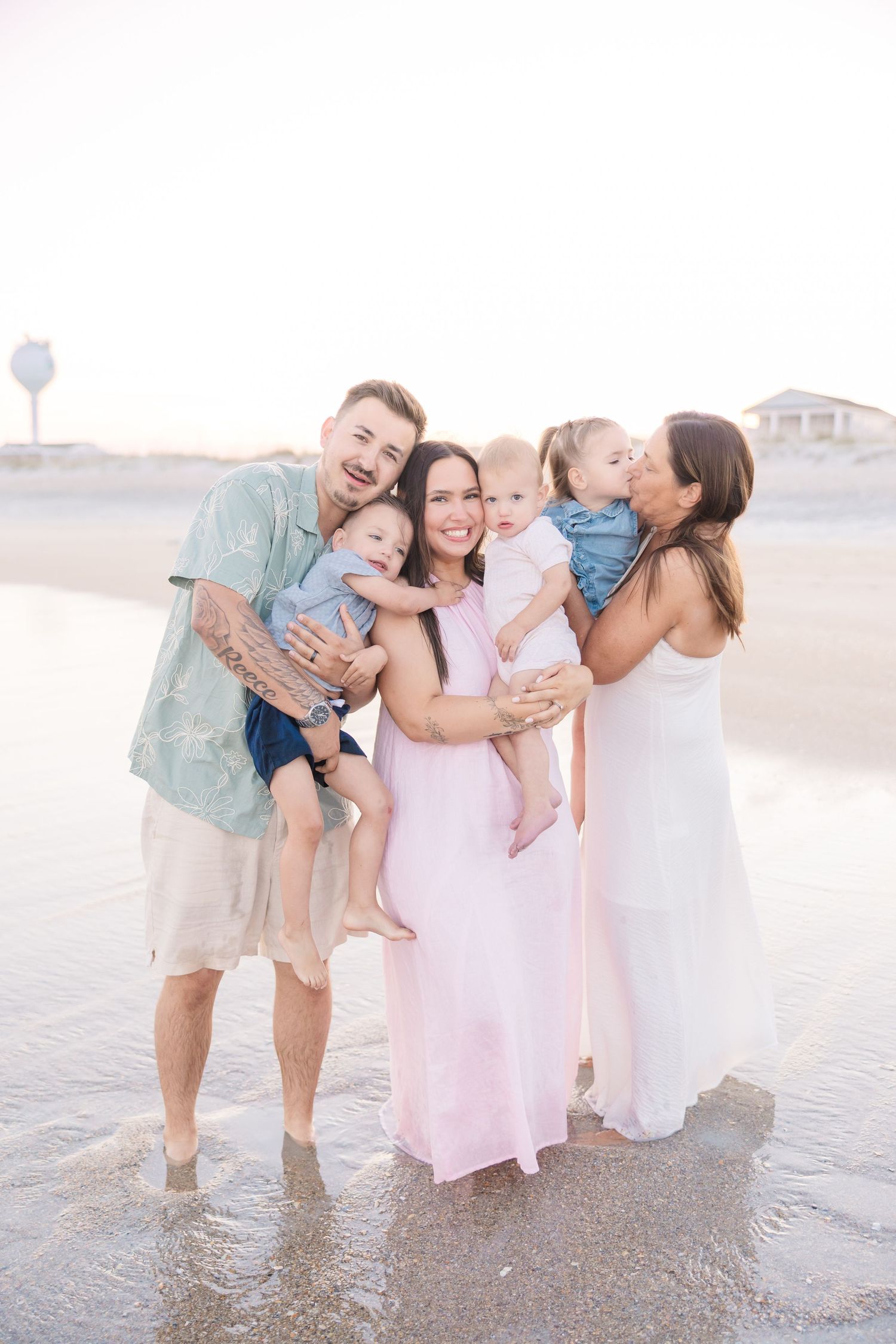 Extended family of five smiles together on beach at sunset, parents holding three young children.