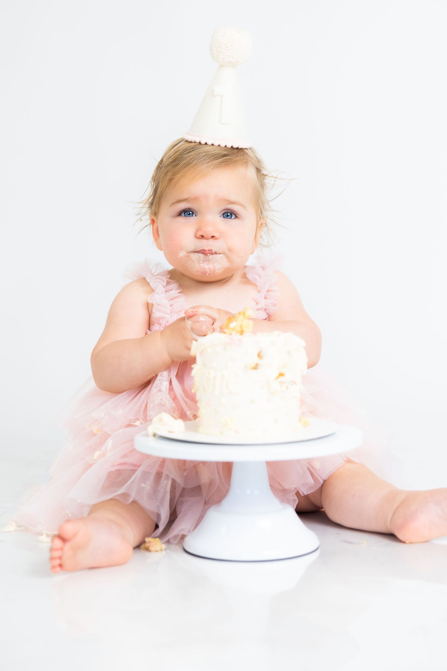Baby in pink dress enjoys cake smash photo session with white frosted cake.