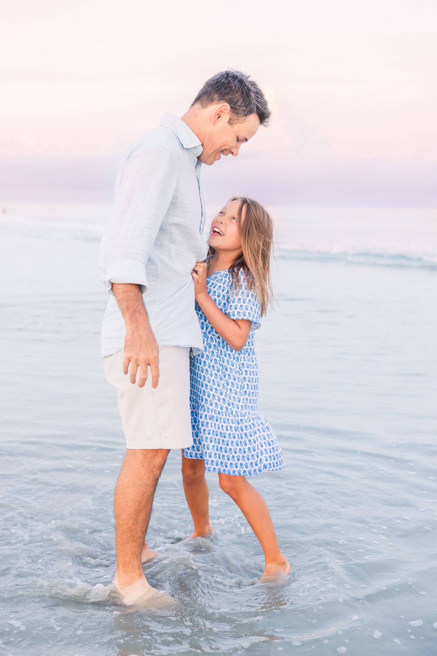 A tender beach moment at sunset with two people standing in shallow ocean water wearing light blue and white outfits.