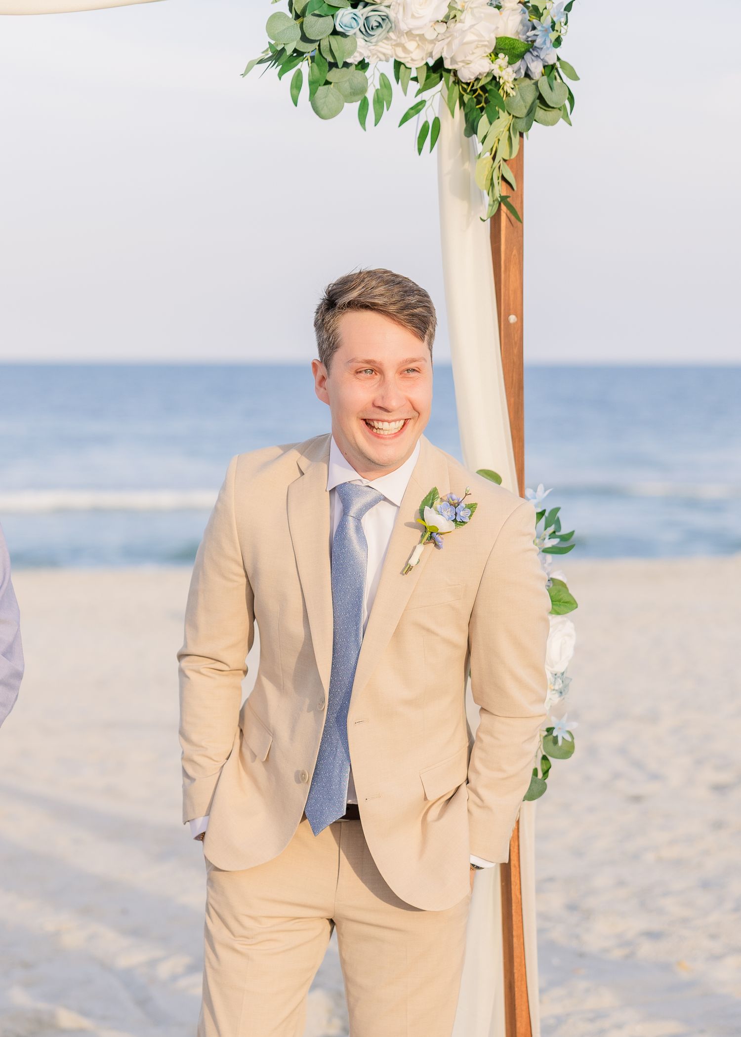 Smiling groom in tan suit with blue tie and boutonniere stands before floral beach wedding arch in Carolina Beach near Wilmington, NC.