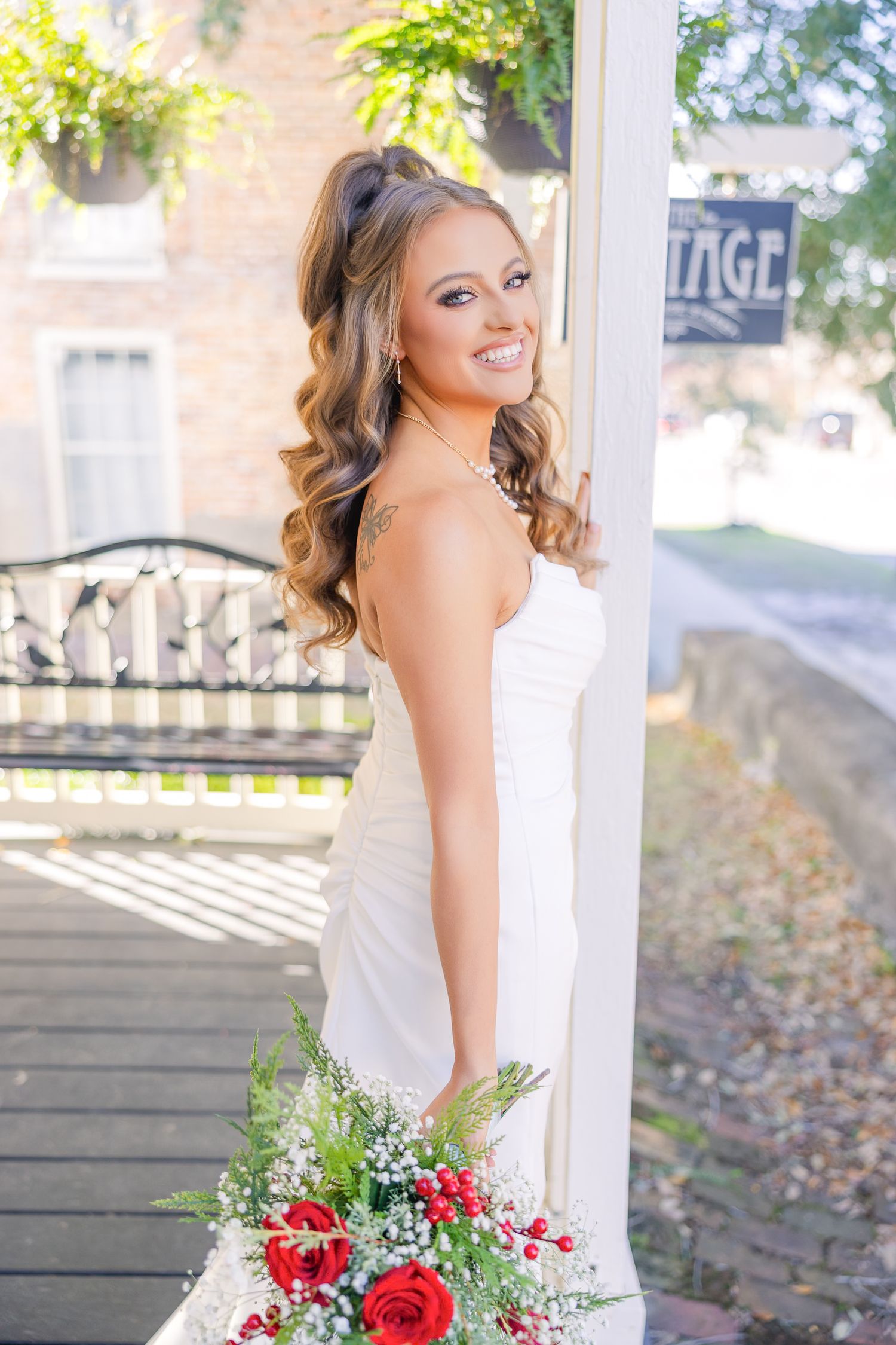 A bride in a white dress poses elegantly against white pillars with red and green floral bouquet.