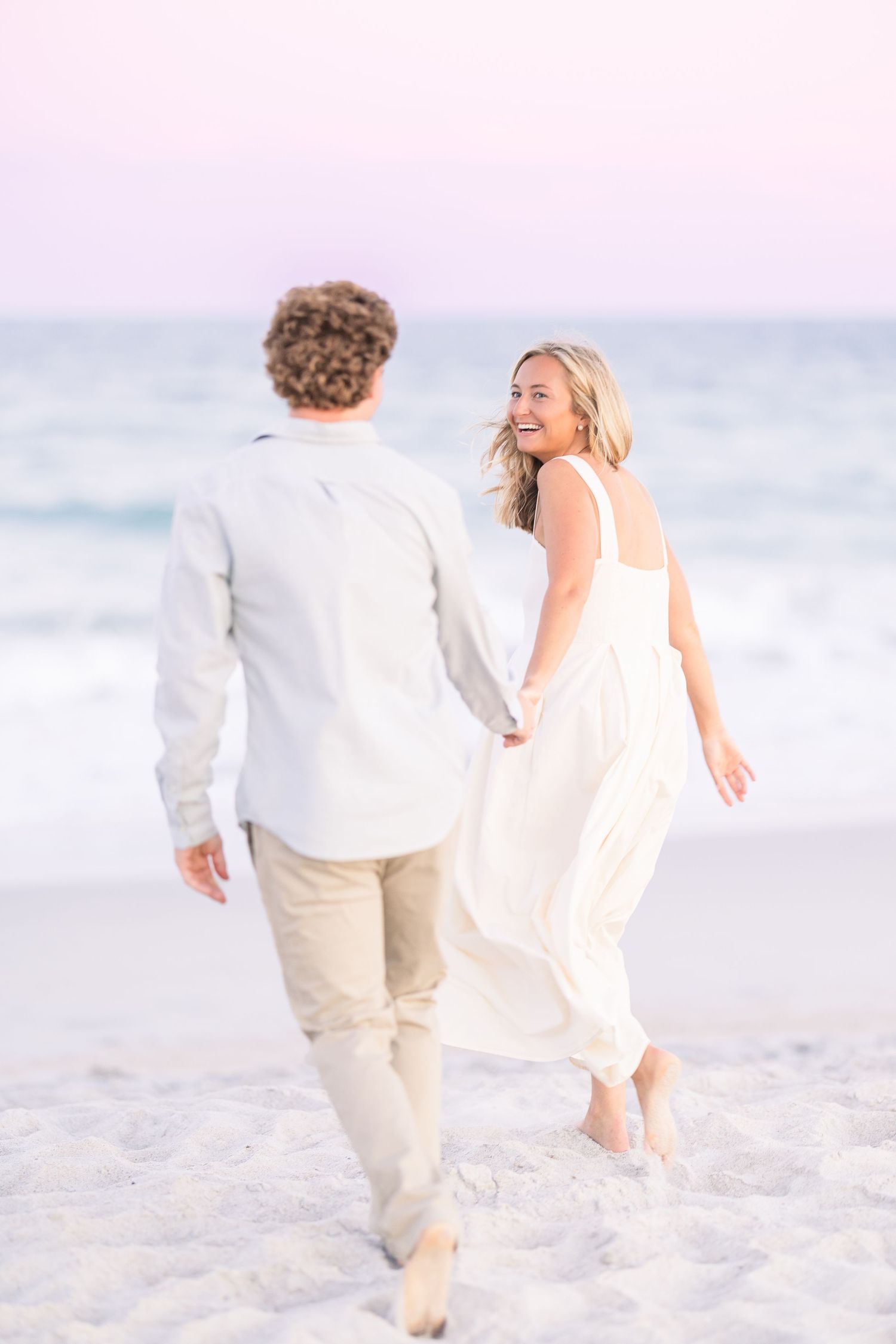 Couple walks along beach shoreline in coordinated white and beige outfits.
