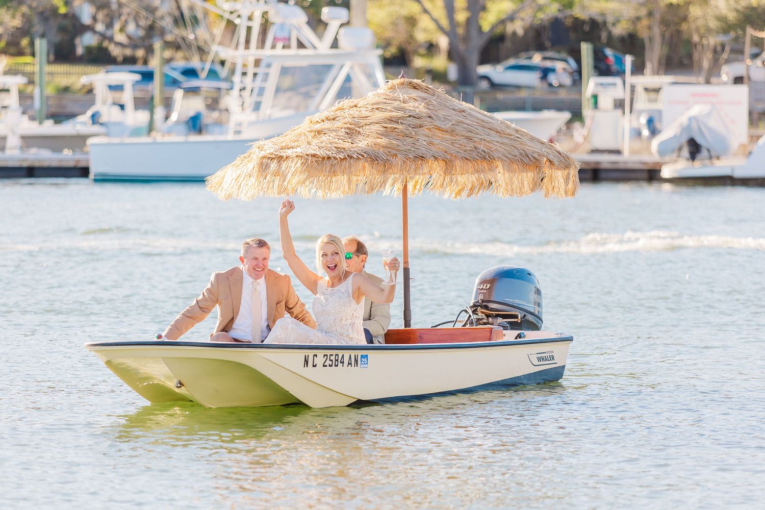 Newlyweds wave from a small white boat decorated with a thatched umbrella landfall wedding bride groom light and airy classic wilmington