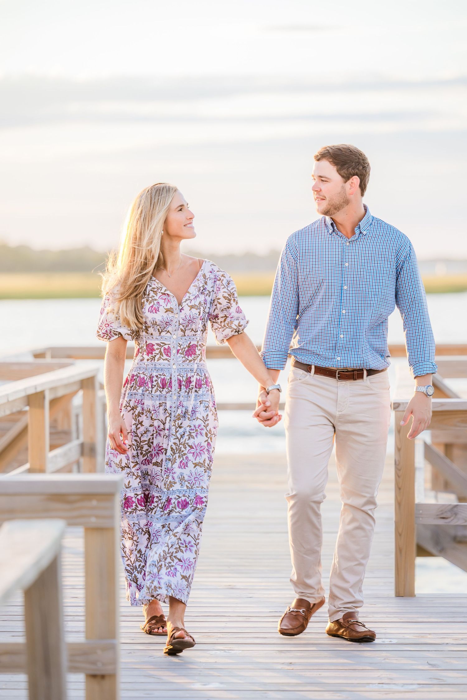 A couple walks hand in hand along a waterfront boardwalk during golden hour.