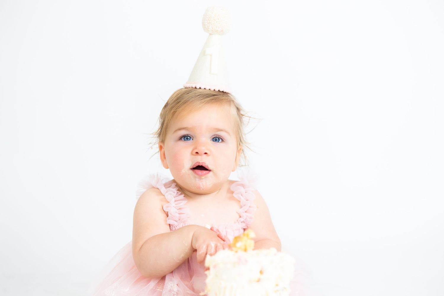 Baby in pink dress and party crown sitting next to white birthday cake during photo session.