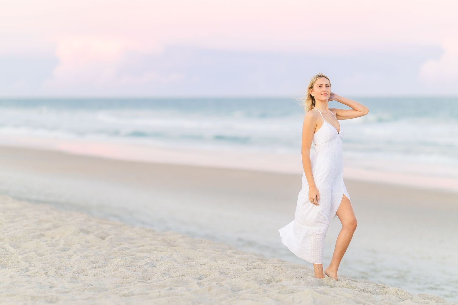 A person in a white dress stands gracefully on sandy beach with soft pink sunset sky.