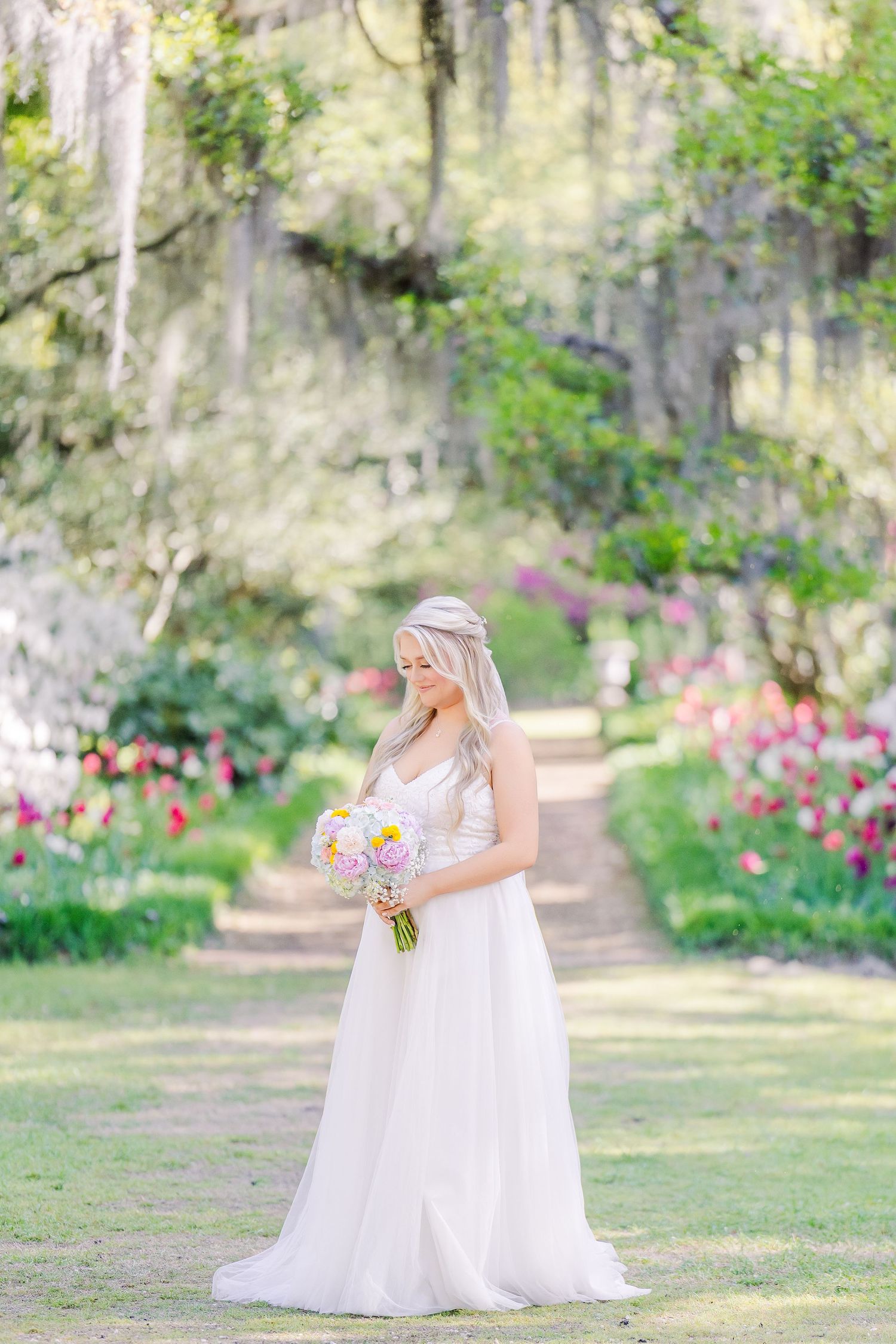 Bride in white gown holds pink bouquet in lush garden path lined with colorful tulips and moss-draped trees in Airlie Gardens in Wilmington,