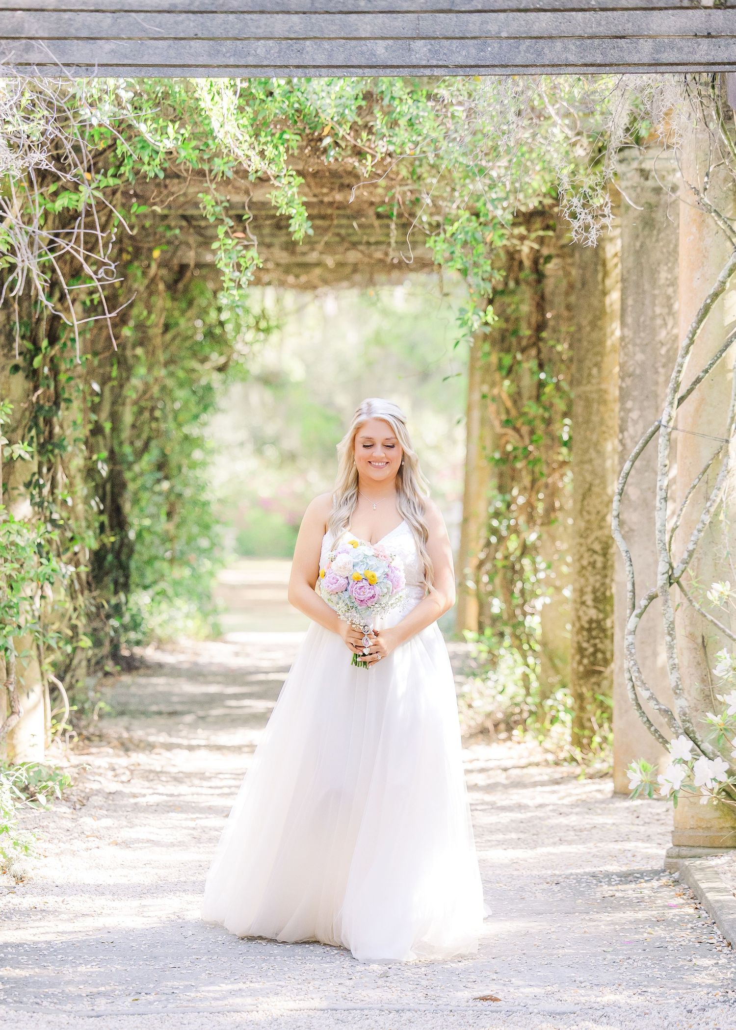 Bride in white ball gown holding colorful bouquet stands under vine-covered pergola in lush garden setting in Airlie Gardens in Wilmington,