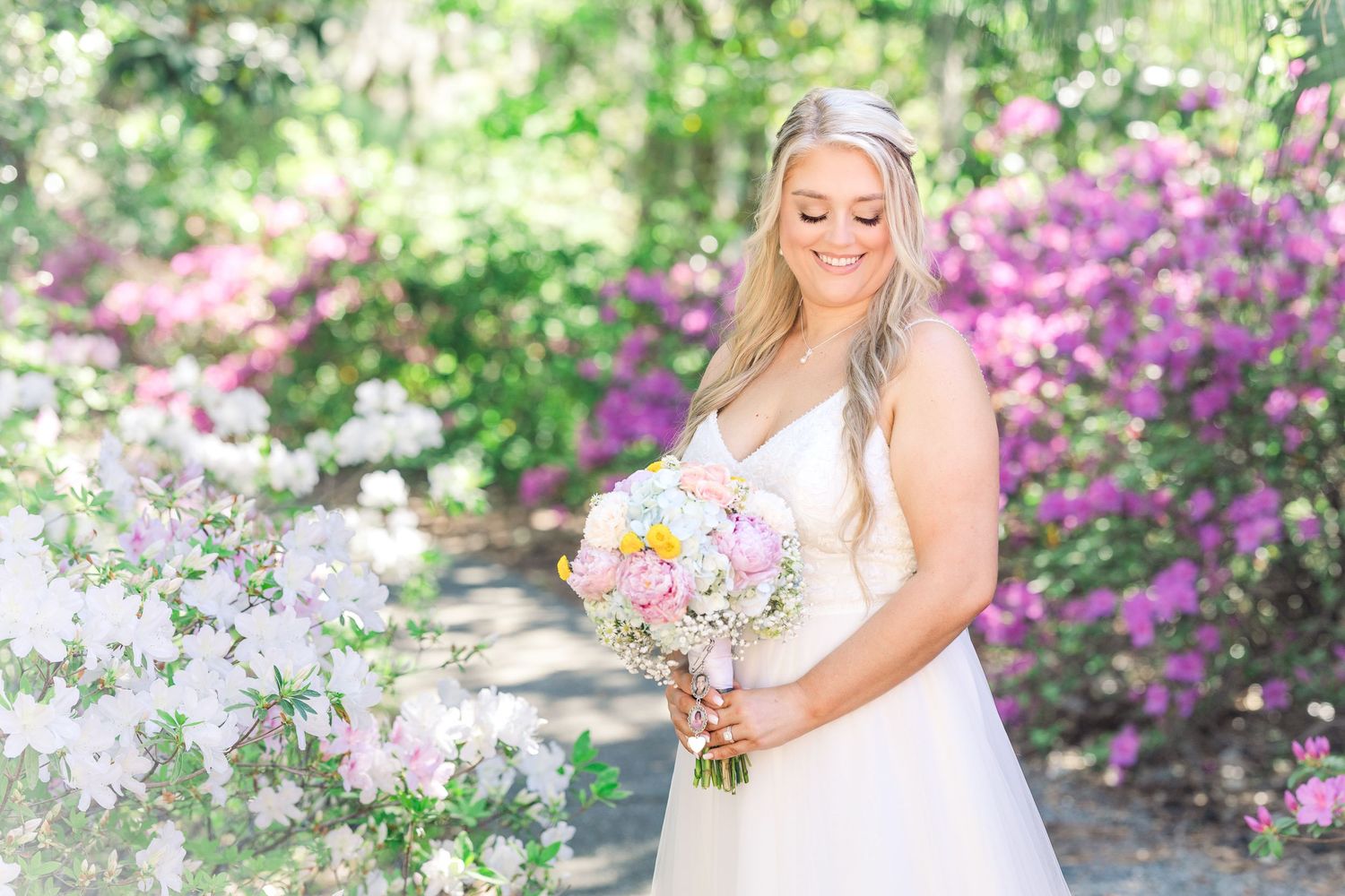 Bride in white gown holding pink bouquet smiling among blooming purple and pink flowers in garden in Airlie Gardens in Wilmington, NC
