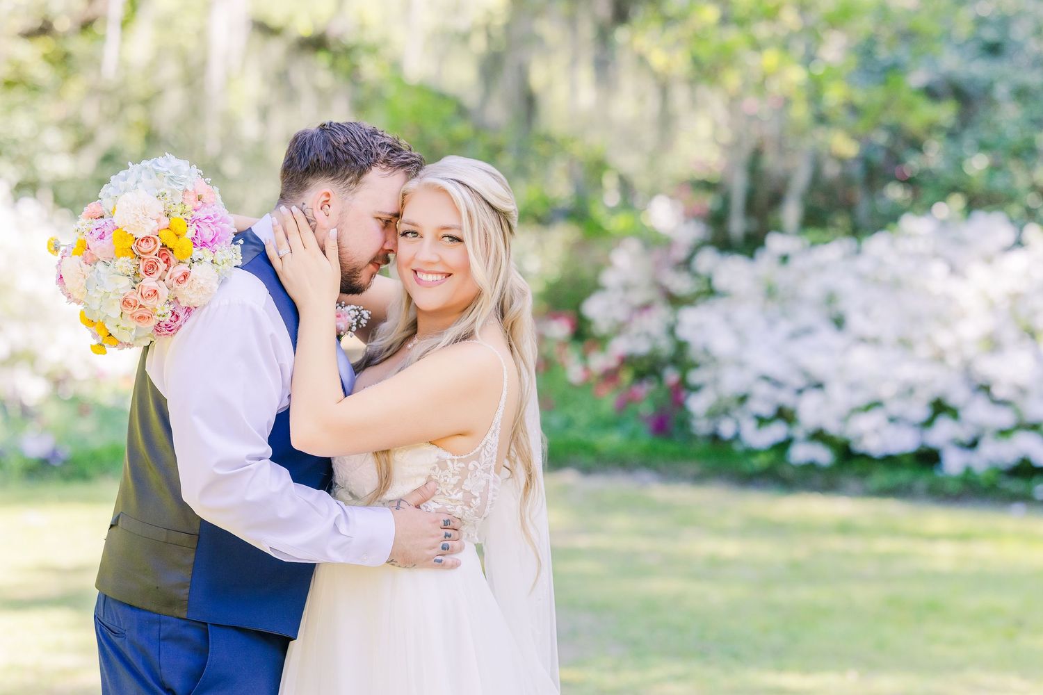 Bride smiling as groom kisses her cheek outdoors, holding colorful bouquet with blooming trees in background.