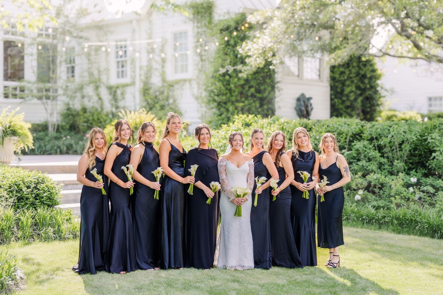 Bride in white gown poses with bridesmaids in navy dresses holding yellow bouquets outside elegant estate.