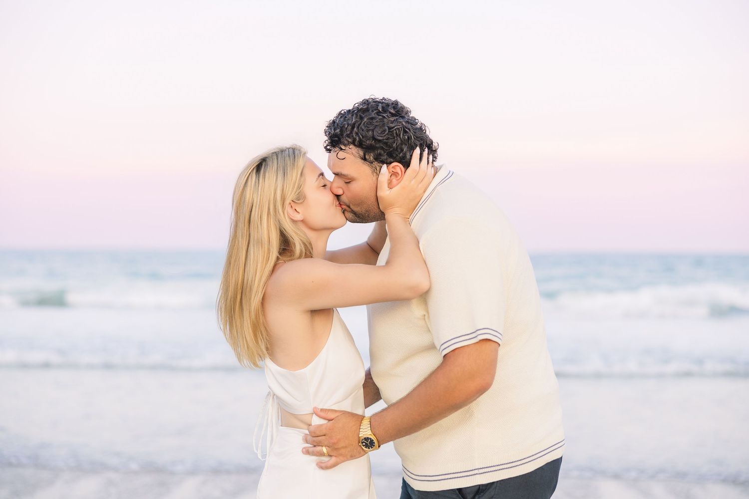 Couple sharing a kiss on the beach at sunset with ocean waves in the background.